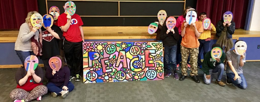 Students with their art masks in front of their faces with their Daniel Cascardo Peace mural in the middle