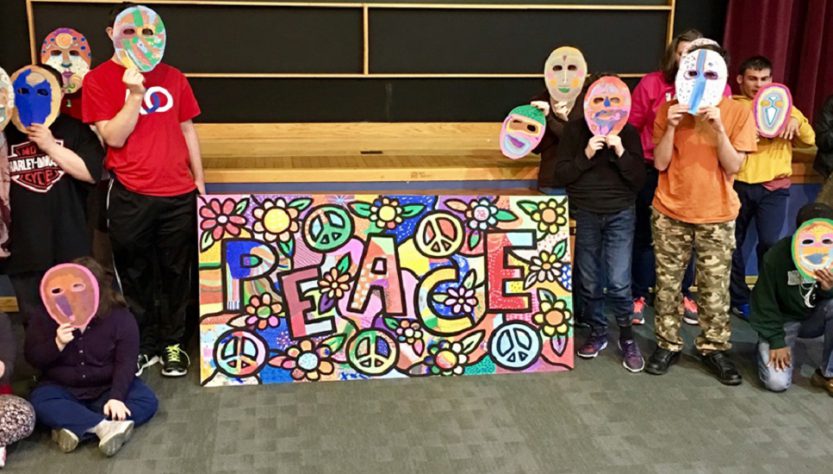 Students with their art masks in front of their faces with their Daniel Cascardo Peace mural in the middle