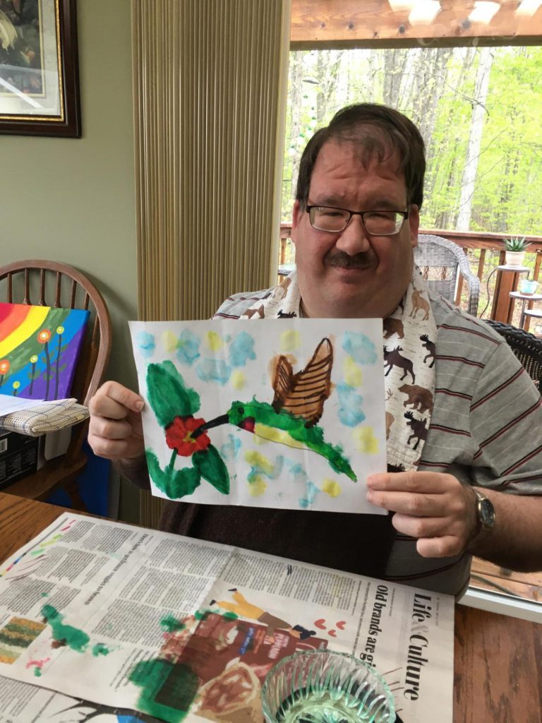 Stephen Broadworth, Club Create Swartz Creek member showing of his hummingbird painting sitting at his kitchen table.