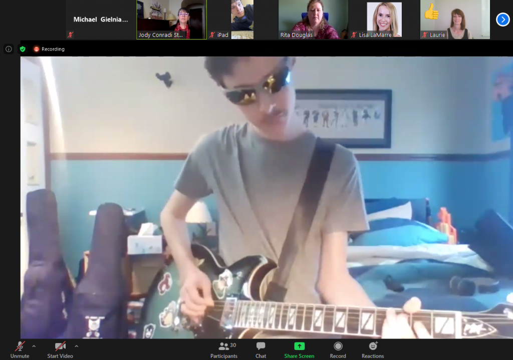 A picture of a client in sunglasses playing electric guitar on camera from his bedroom.