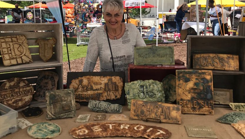 Clay artist standing in her art fair booth with Detroit inspired clay artwork displayed on the table in front of her