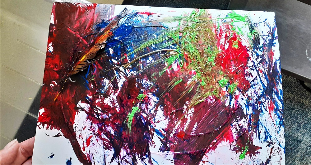 Red, blue green and purple abstract painting on a white canvas with a feather embedded in the painted work.