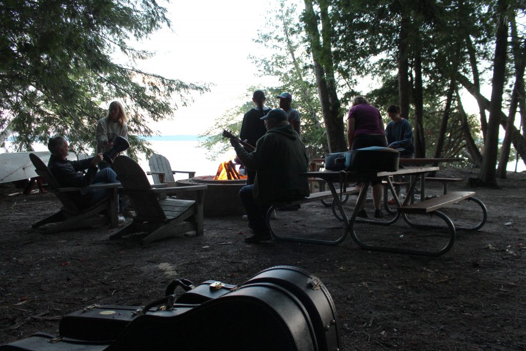 Attendees sitting around the fire by Higgins Lake, playing music on the guitar and drums. 