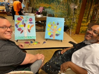 Michael and Rico looking back at photographer and smiling with their paintings of trees on the tabletop easels in front of them