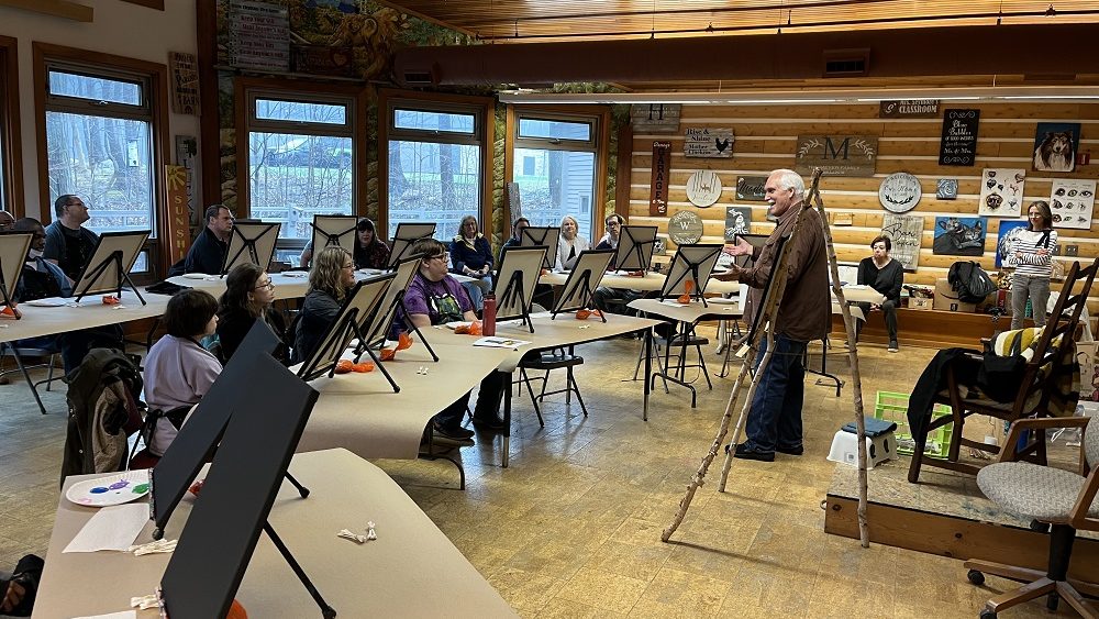 Robert White standing in front of members of Club Create North Oakland County who are sitting in front of tabletop easels getting ready to paint.