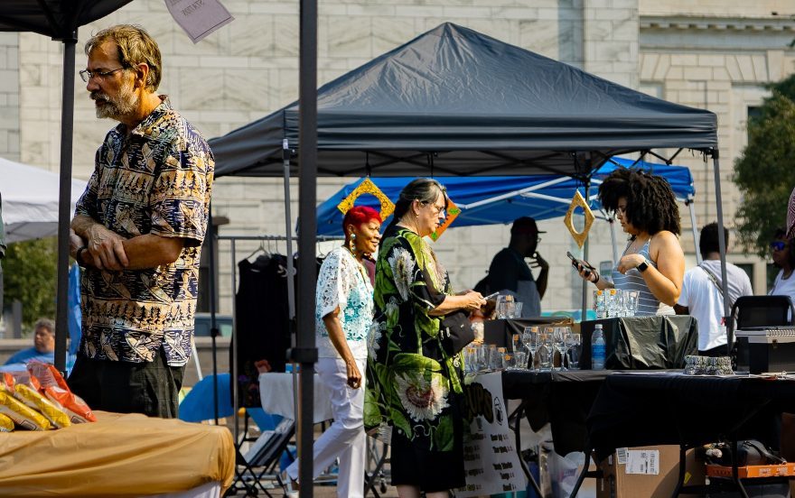 two women at an artist booth, one making a purchase, with a man in the foreground at a different booth and another artist booth behind the two women